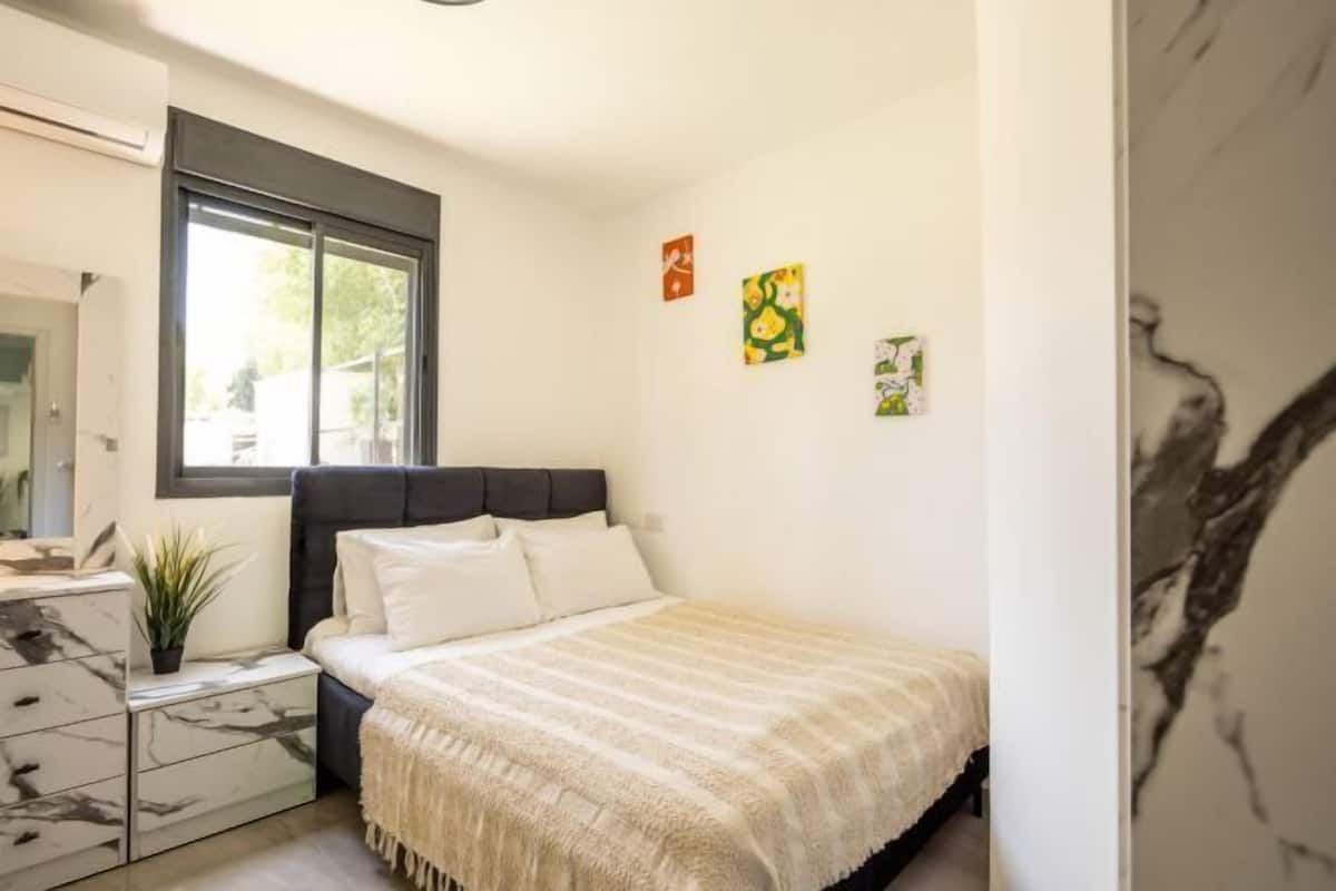 A bedroom with a bed, a nightstand, a dresser, a window, and wall art.