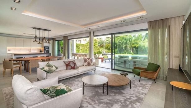 A modern living room with a sofa, coffee table, and chairs, a dining area with a table and chairs, and a kitchen with a counter and hanging lights. - All About Villa Gardenias (Marbella)