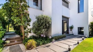 A modern building with a clean, white exterior, a stone pathway leading to the entrance, and lush greenery around. - All About Villa Gardenias (Marbella)