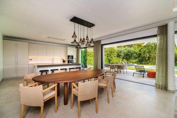 Une cuisine moderne dotée d’un grand îlot central, d’un coin repas avec des chaises et offrant une vue sur la verdure environnante.