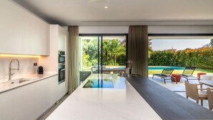A modern kitchen with a large island, a dining area with chairs, and a view of a swimming pool. - All About Villa Gardenias (Marbella)