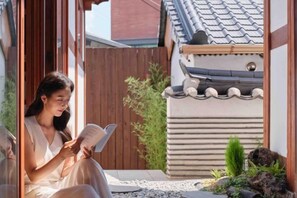 Une personne est assise sur un banc, lisant un livre dans une cour bordée d’une clôture en bois, surmontée d’un toit en tuiles, et agrémentée de quelques plantes.