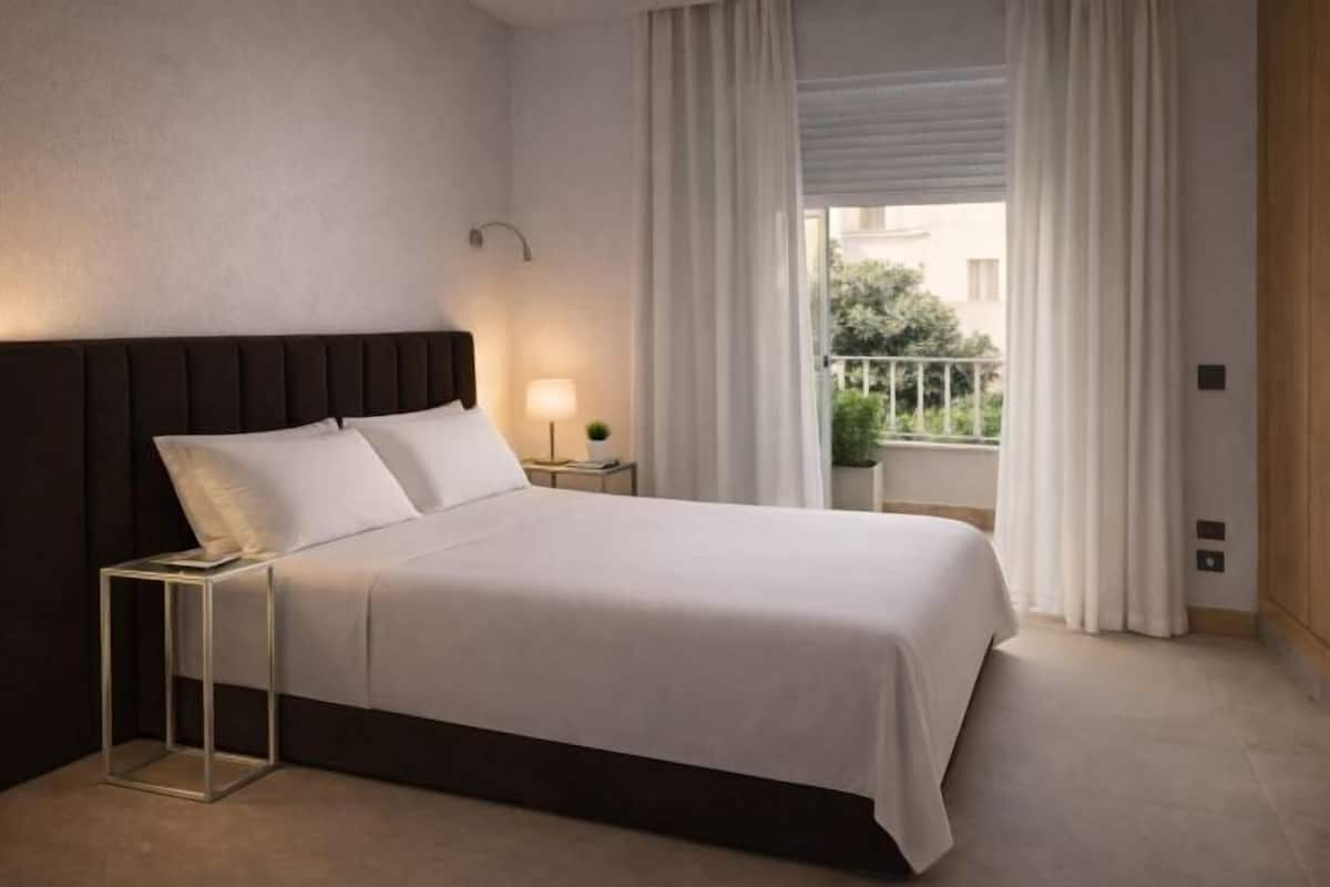Ein Hotelzimmer mit Bett, Nachttisch, Lampe, einem Fenster mit Vorhängen und Blick auf Grünfläche.