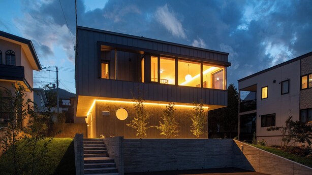 Une maison moderne à deux étages, dotée d’une grande baie vitrée à l’étage supérieur, d’une fenêtre circulaire à l’étage inférieur et d’un escalier menant à l’entrée.