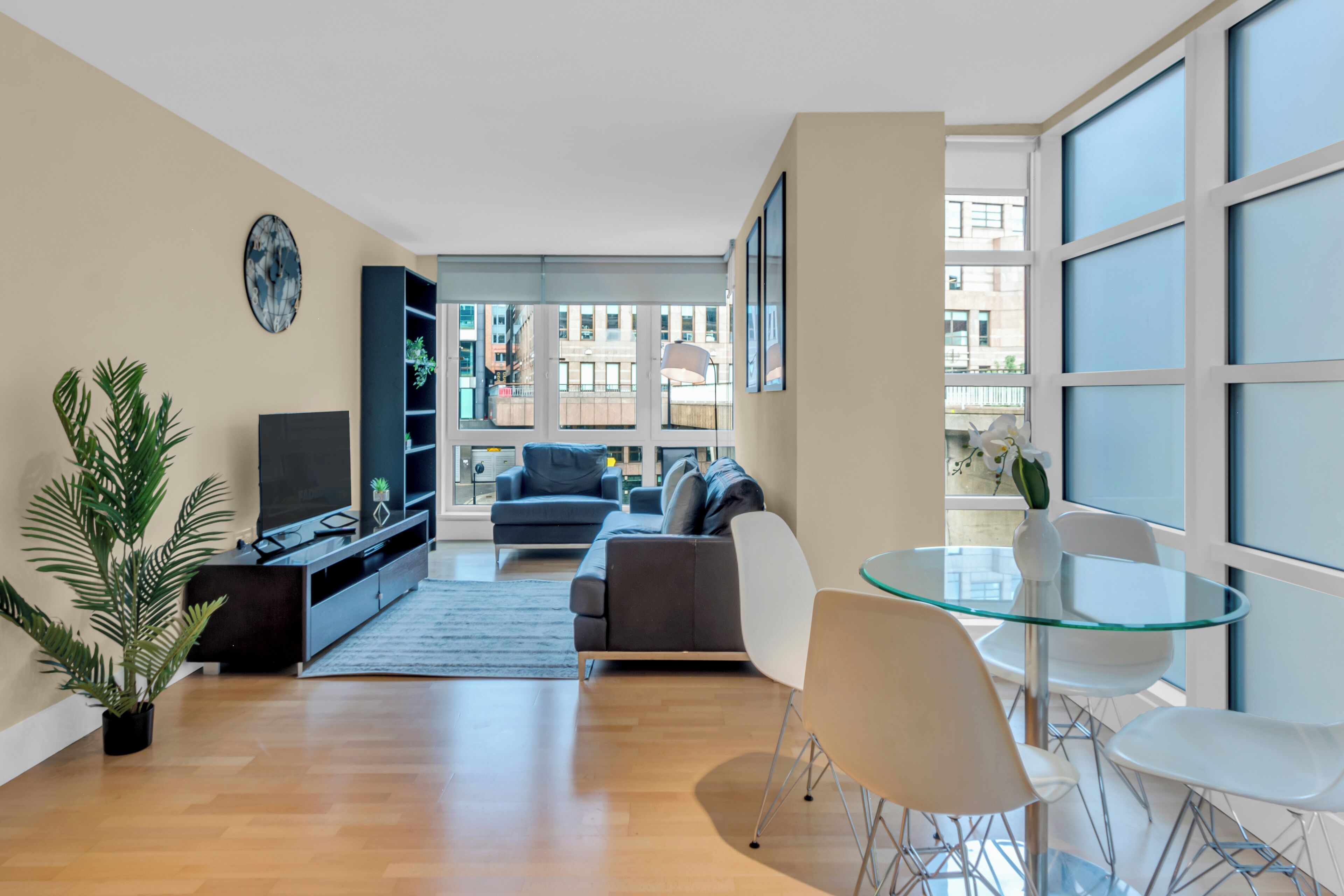 Una sala de estar moderna con sofá, televisor, mesa de vidrio, sillas y un amplio ventanal con vistas a la ciudad.