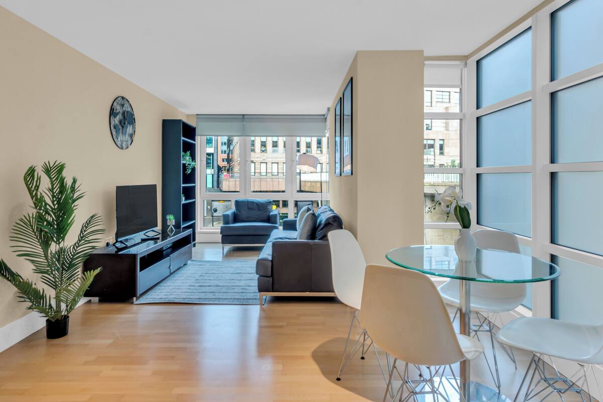 Un salon moderne comprenant un canapé, un téléviseur, une table en verre, des fauteuils et une grande fenêtre offrant une vue sur la ville.