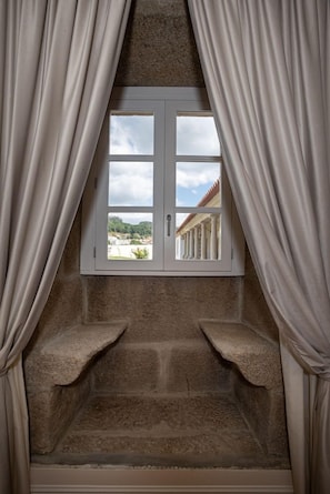 Una habitación con ventana enmarcada en piedra, cortinas y vista a un edificio con columnas.