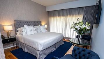 Una habitación de hotel con una cama grande, una mesita de noche con lámpara, una mesa redonda con un jarrón de flores y un sillón azul.