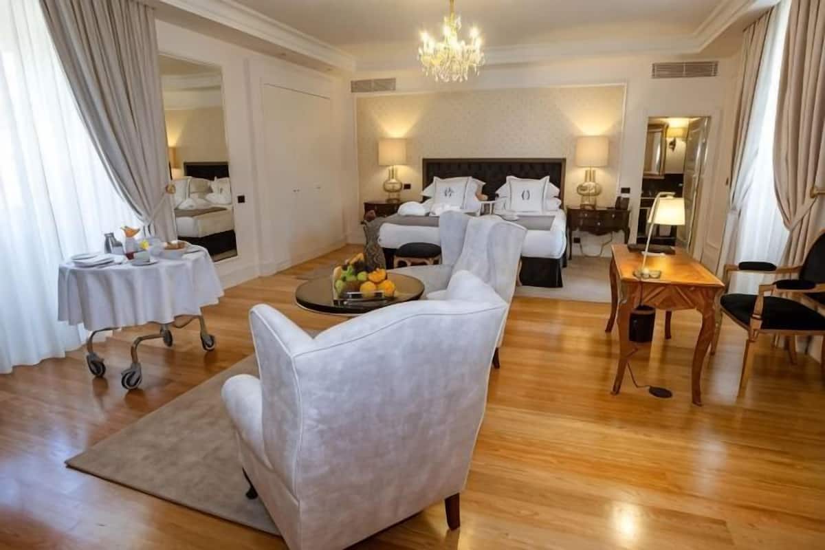 Quarto de hotel com cama, área de estar com sofá e poltronas, uma mesinha com uma cesta de frutas e um lustre.