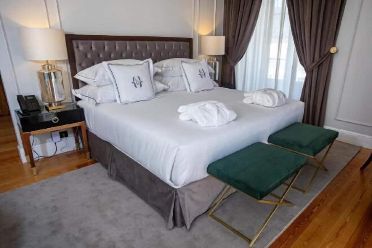 Uma cama bem arrumada com lençóis e almofadas brancos, uma mesa de cabeceira com abajur e um banco verde junto à janela.