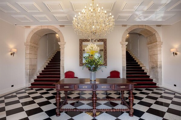 Una imponente escalera con candelabro, piso a cuadros y una mesa con sillas.