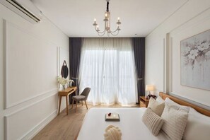 Habitación de hotel con una cama grande, un escritorio, una silla, una araña de cristal y un ventanal con cortinas.