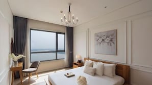 A hotel room with a large bed, a desk, a chair, a nightstand, a chandelier, a window with a view, and a framed artwork on the wall. - Diamond Island Highfloor RiverView 8min to D1 (Ho Chi Minh City)