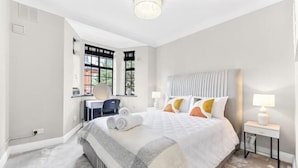 A hotel room with a large bed, a desk with a chair, a window with blinds, and a bedside table with a lamp. - Stylish 3-Bedroom Flat in Belsize Park – Perfect for Families & Business Trips! (London)