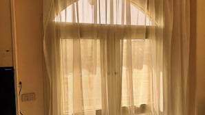Quarto com janela coberta por cortinas leves, tomada visível e interruptor de parede.