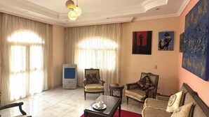 Uma sala de estar com sofá, poltronas, uma mesa pequena e uma televisão.