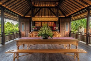 A wooden dining table with a potted plant centerpiece, surrounded by chairs, under a thatched roof structure. - Villa Kapungkur (Bali)