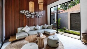 A modern living room with a sectional sofa, a coffee table, and a round ottoman. There are two hanging light fixtures and a decorative wall piece. - Villa Surya by BaliSuperHost (Bali)