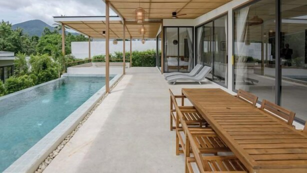 A modern outdoor pool area with a wooden table and chairs, glass doors, and a view of greenery and mountains. - Tropical Peaceful Poolside Retreat by Sea N' Rent (Koh Phangan)