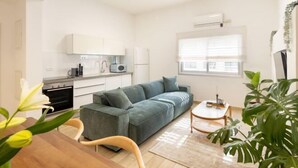 Un salon moderne avec un canapé gris, une table à manger en bois et une petite table basse ronde. On aperçoit un coin cuisine avec des meubles blancs et un four à micro-ondes. Une fenêtre avec des stores est visible, et une plante en pot se trouve au premier plan.