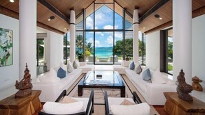 A modern interior with a large glass window, white sofas, a coffee table, and wooden columns. - Luxury Beachfront Villas & Resort (Phuket)