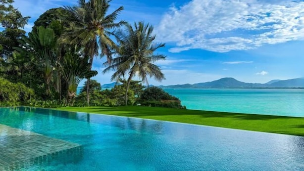An infinity pool with clear blue water, surrounded by lush greenery and palm trees, overlooking a turquoise sea and distant mountains. - Luxury Beachfront Villas & Resort (Phuket)
