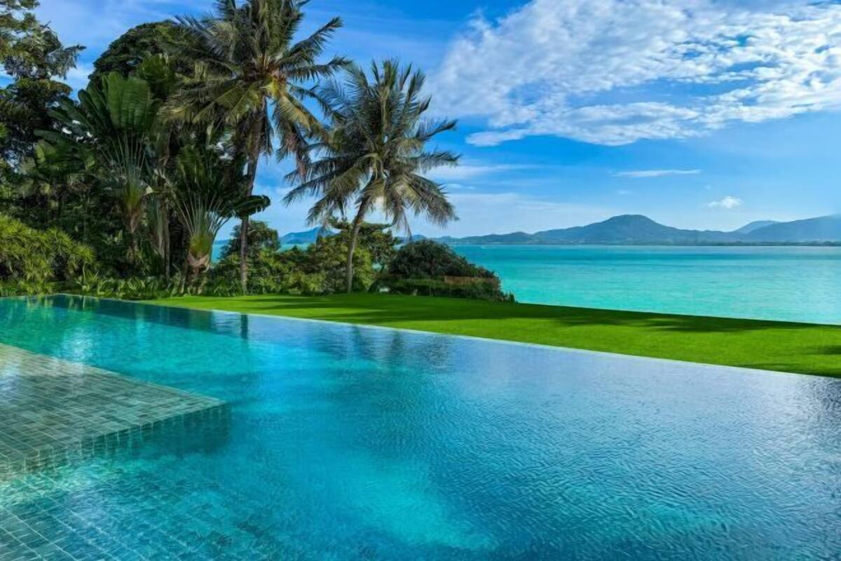 An infinity pool with clear blue water, surrounded by lush greenery and palm trees, overlooking a turquoise sea and distant mountains.