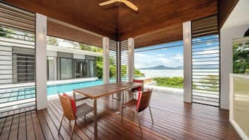 A covered outdoor area with a wooden deck, a table with chairs, and a view of a swimming pool and landscape.