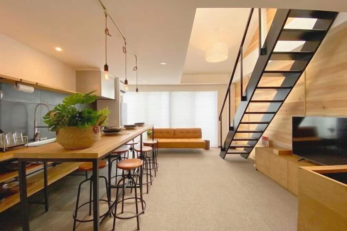 A modern kitchen with a wooden countertop, bar stools, a large plant, and a staircase with a wooden railing.