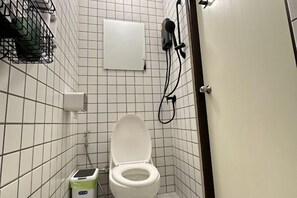 A small, tiled bathroom with a toilet, a trash bin, and a hairdryer mounted on the wall. - Twenty-one Bkk House (Bangkok)