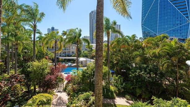 Un hôtel doté d’une piscine entourée d’une végétation luxuriante et de palmiers, avec des bâtiments modernes en arrière-plan et un ciel bleu dégagé.