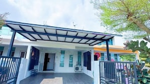 Un espacio al aire libre techado con suelo de baldosas, baranda de metal, y un edificio con fachada blanca y detalles azules al fondo.