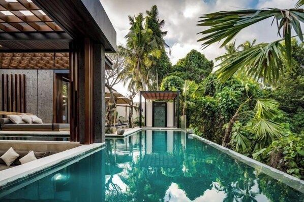 Un espace piscine moderne doté d’une piscine à eau claire, d’une terrasse en bois et d’une végétation tropicale luxuriante.