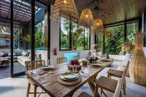 A dining area with a wooden table set for four, surrounded by wicker chairs, and large windows offering a view of the outdoors. - Villa Golden Lion by BaliSuperHost (Bali)