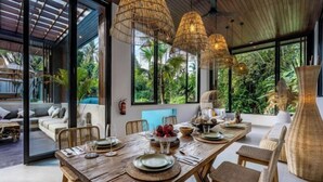 A dining area with a wooden table set for a meal, surrounded by wicker chairs, with large windows offering a view of lush greenery outside.