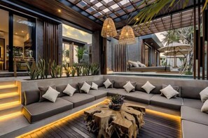 A modern outdoor seating area with a wooden coffee table, grey sofas with white cushions, and a pergola with hanging lights.