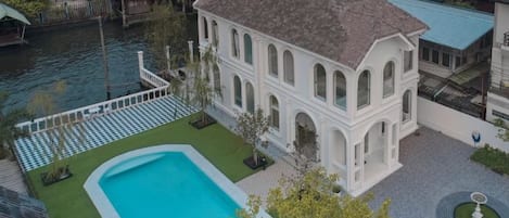 Un edificio blanco con piscina, rodeado de un jardín y una masa de agua.