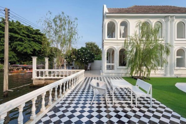 A checkered terrace with white chairs, a white building with arches, and a river in the background. - Arpo Pool Villa Riverside Bangkok (Bangkok)