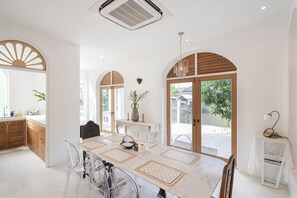A modern kitchen with white cabinets, a dining table with placemats, and a view of the outdoors through large windows. - Arpo Pool Villa Riverside Bangkok (Bangkok)
