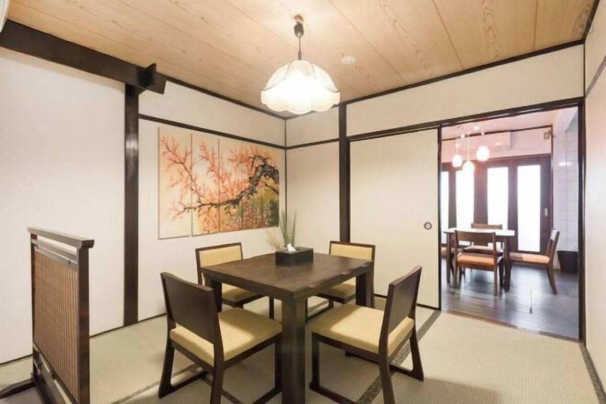 A traditional Japanese-style room with tatami flooring, a dark wooden table with chairs, a hanging light fixture, and a painting on the wall.