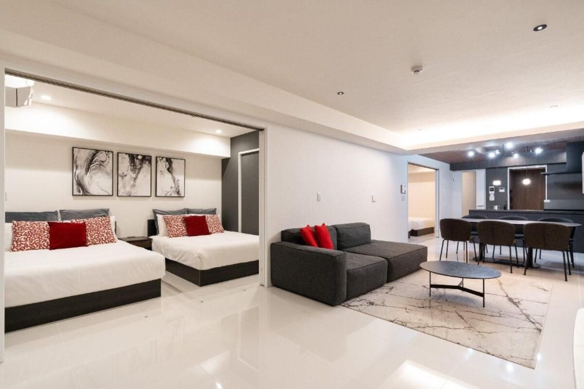 A modern hotel room with two beds, a sofa, a dining area, and a kitchen.