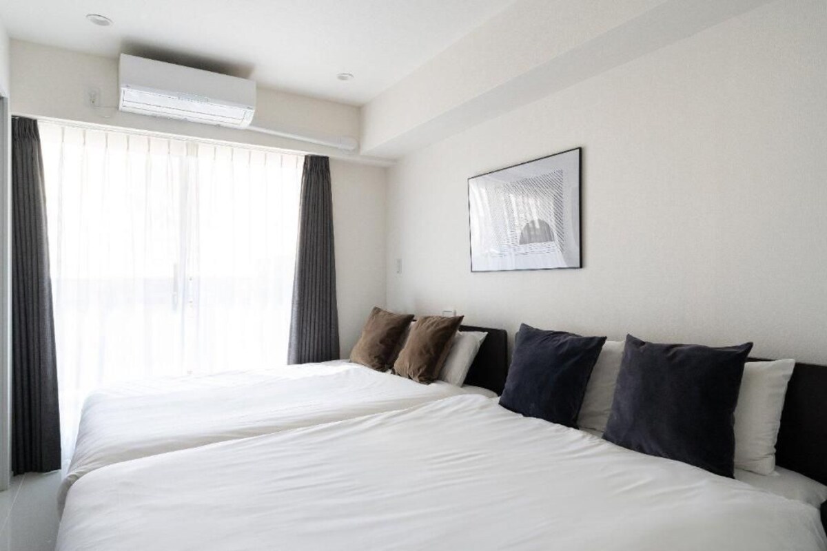 A hotel room with two beds, a large window with curtains, an air conditioning unit, and a framed picture on the wall.