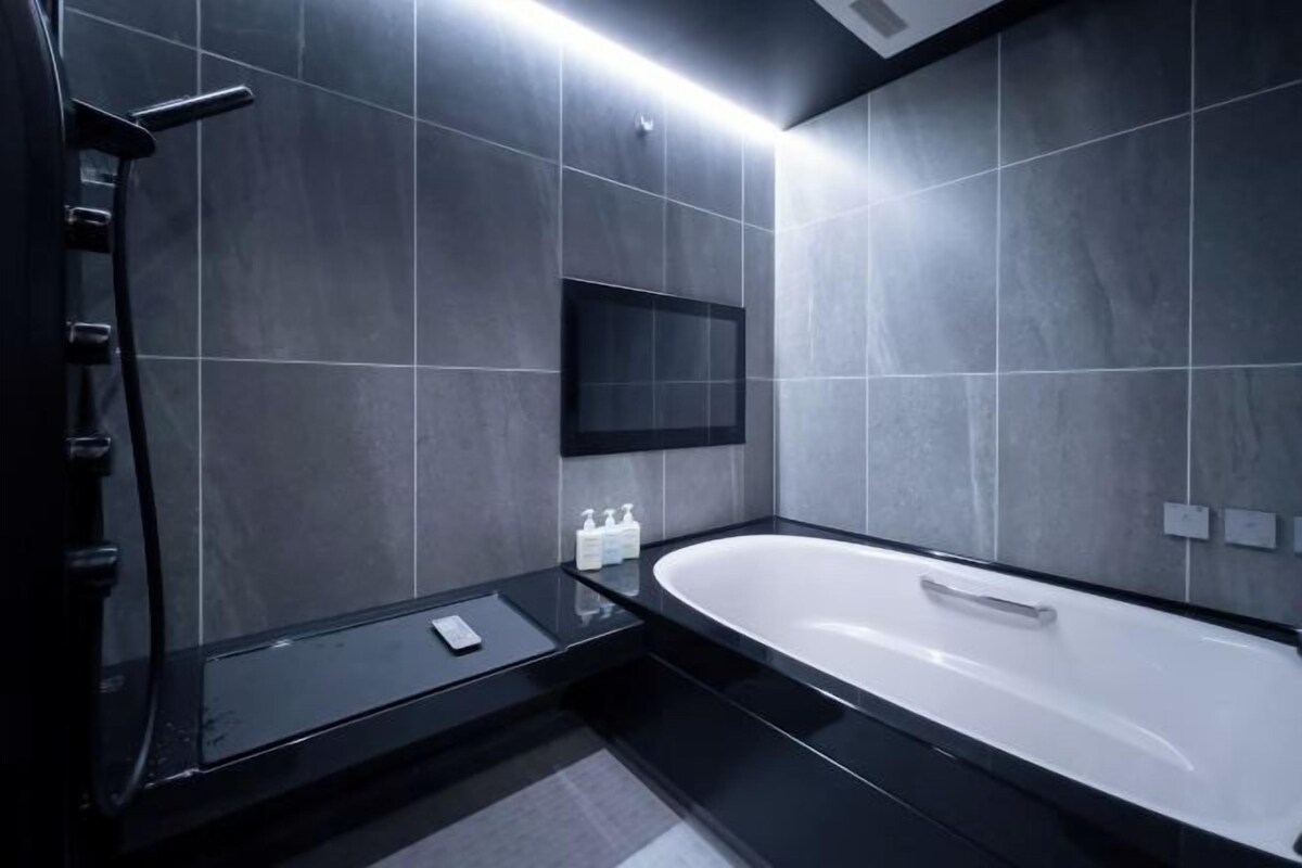 A modern bathroom with a large bathtub, dark tiled walls, a black countertop, and a mounted TV.