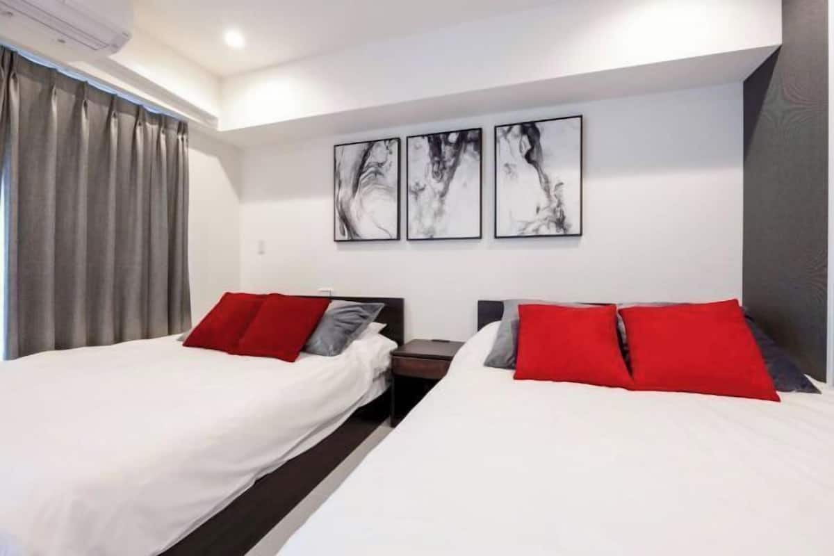 A hotel room with two beds, each with red and grey pillows, and a nightstand between them. There are three abstract paintings on the wall above the beds.