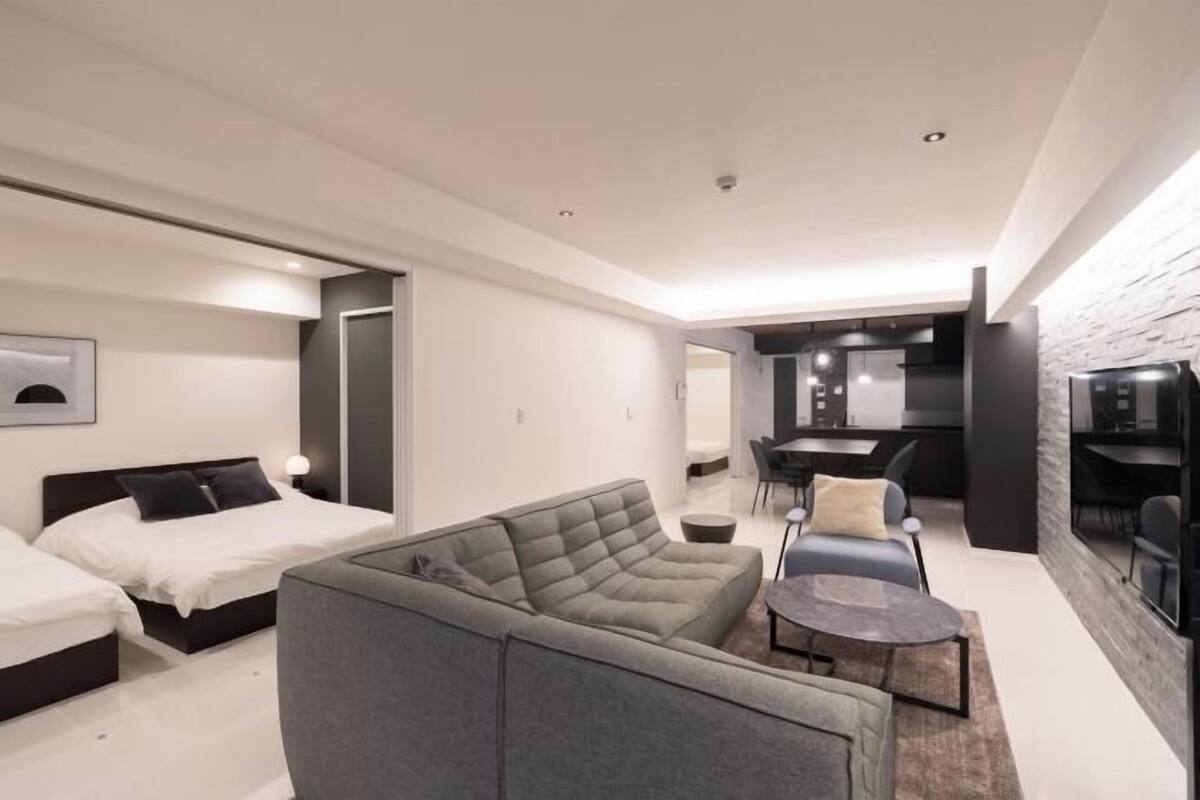 A modern hotel room with a sofa, two beds, a dining area, and a kitchen.