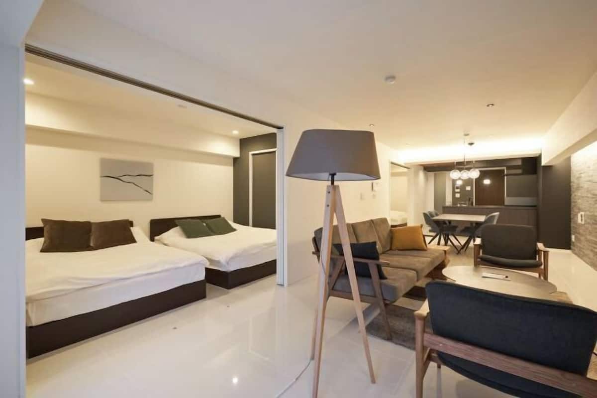 A modern hotel room with two beds, a sofa, a dining table, and a lamp.