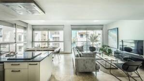 A modern kitchen with a large island, a dining area with a sofa and chairs, and a TV on the right. - Chic 3BR Apt with Balcony, Mamad (Tel Aviv)