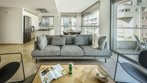 A modern living room with a grey sofa, a coffee table with a magazine, and a view of a cityscape through large windows. - Chic 3BR Apt with Balcony, Mamad (Tel Aviv)