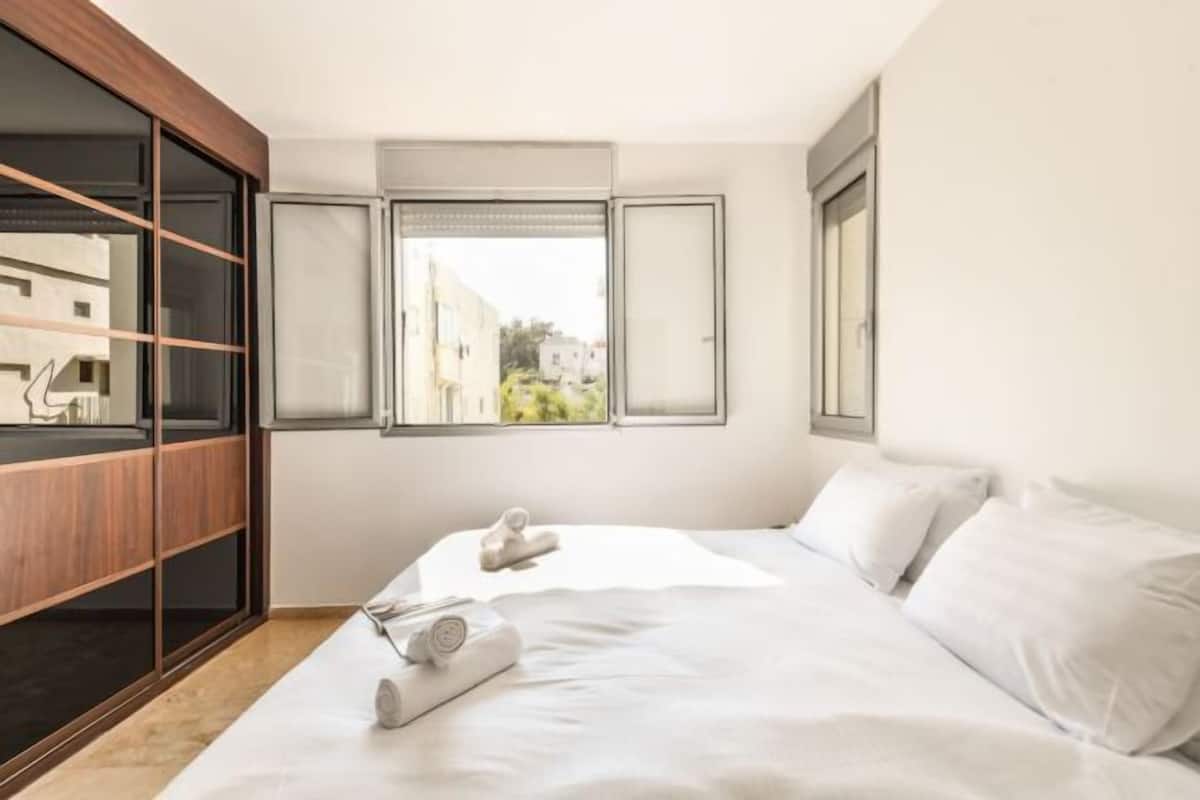 A bedroom with a bed, a wooden wardrobe, and a window with a view of buildings.