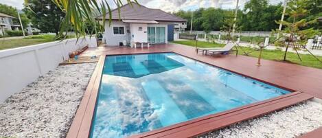 A rectangular swimming pool with a wooden deck, surrounded by a white wall and a gravel area.